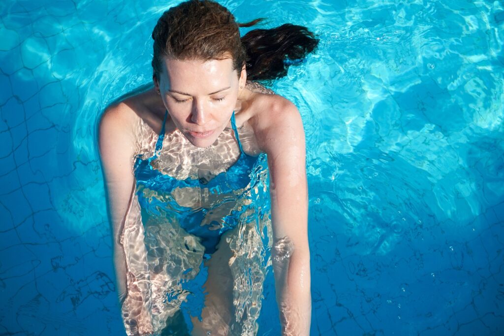 Ruskeahiuksinen nainen vesijumppaa altaan kaiteella sinisessä uima-asussa.