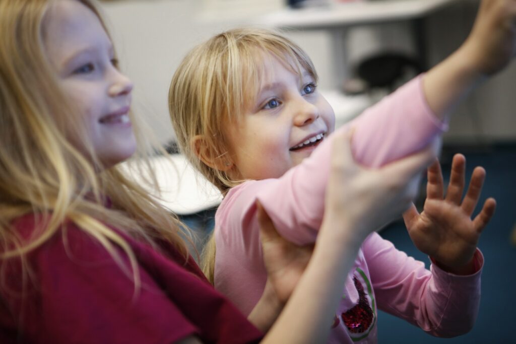 Kaksi vaaleahiuksista alakouluikäistä tyttöä innoissaan kädet ojossa laittamassa jotakin seinälle tai johonkin esineeseen jotain, joka rajautuu kuvan ulkopuolelle.