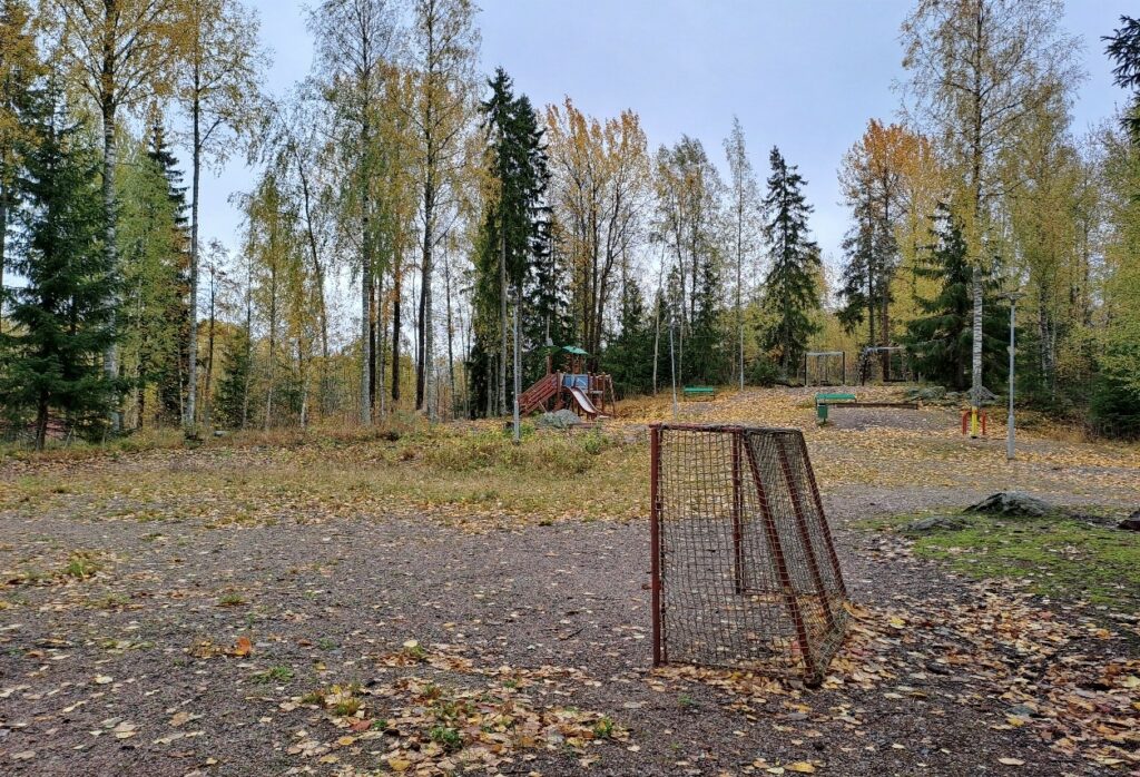 Hiekkakentällä pallopelien maali ja syksyisiä lehtiä. Taustalla leikkikentän liukumäki. Kenttää ympäröi puut.