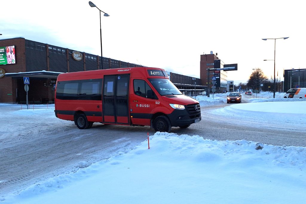 Punainen pikkubussi talvisella tiellä, iso matala rakennus taustalla.