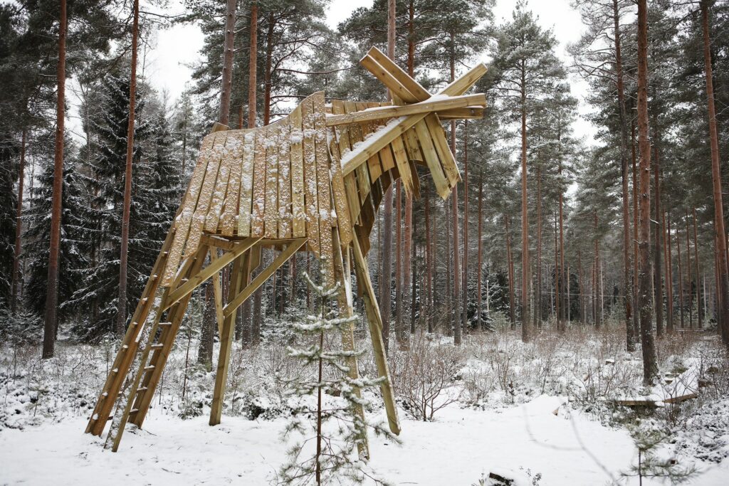 Puusta rakennettu suuri hirvi lumisen maiseman keskellä, taustalla metsää.