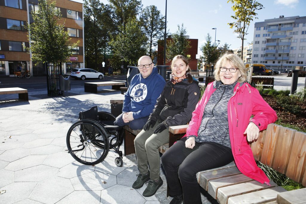 Kaksi naista ja yksi mies istuvat yhdessä aukiolla penkillä, hymyilevät kameralle. Miehen vieressä on pyörätuoli.