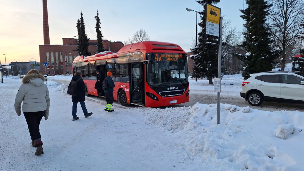 Lumisessa kaupunkimaisemassa punainen bussi pysäkillä seisahtuneena ja ihmisiä nousemassa kyytiin.