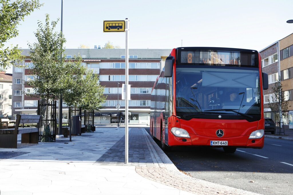 Punainen bussi pysähtyneenä pysäkille. Pysäkin kohdalla menee pinnastaan uusittu kevyen liikenteen väylä. Puissa on lehtiä, pilvetön taivas.