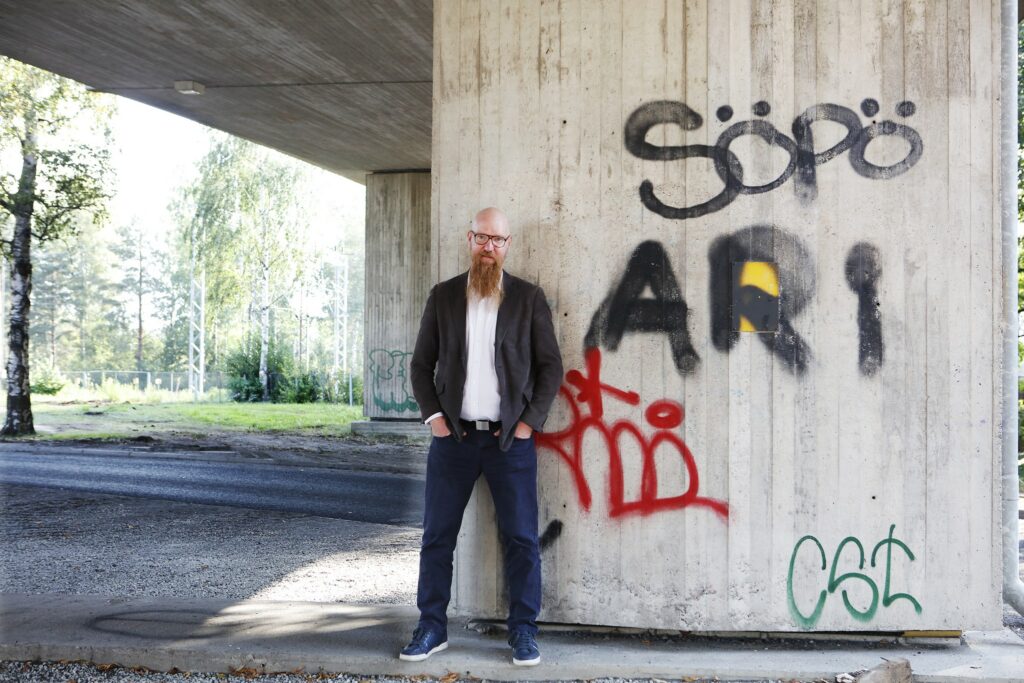 Mustatakkinen, parrakas mies suuren sillan alla. Sillasta näkyy kuvassa betoninen jalka, jossa graffiti "Söpö Ari". Mies seisoo tekstin vieressä.