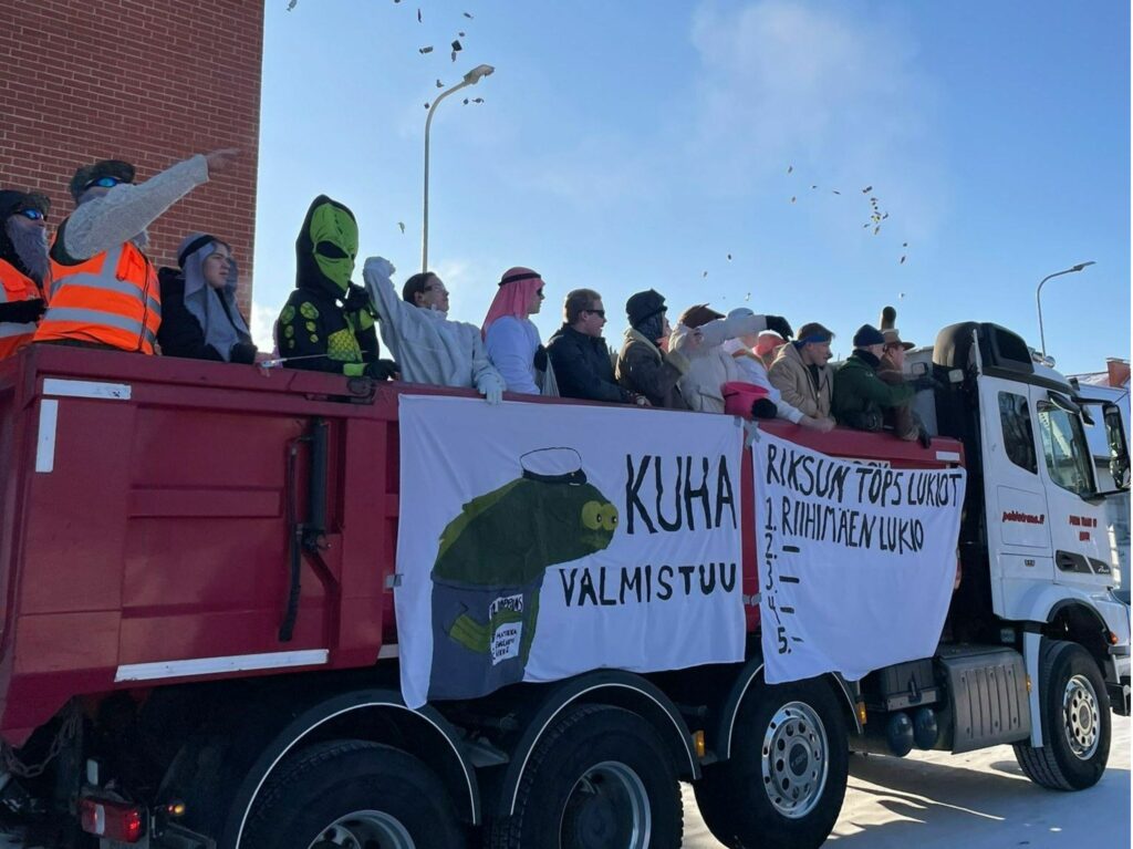 Rekka, jonka kyydissä nuoria pukeutuneina naamiaisasuihin. Rekan kyljessä roikkuu kaksi lakanaa, joista toisessa lukee Kuha valmistuu sekä maalattu kuhan kuva ja toisessa lakanassa lukee teksti Riksun TOP 5 lukiot. Ykkössijalla on Riksun lukio ja loput sijat ovat tyhjät. Karkkeja ilmassa.