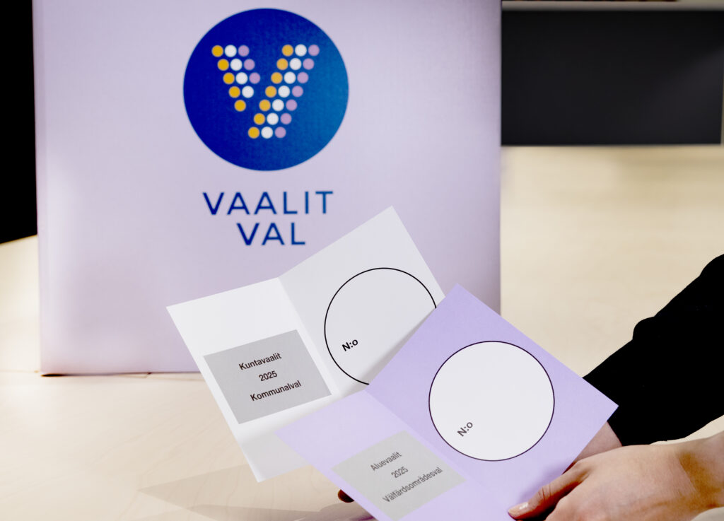 Kädet, jotka pitelevät kahta lipuketta, valkoista ja violettia. Taustalla äänestyslaatikko, jossa lukee Vaalit.